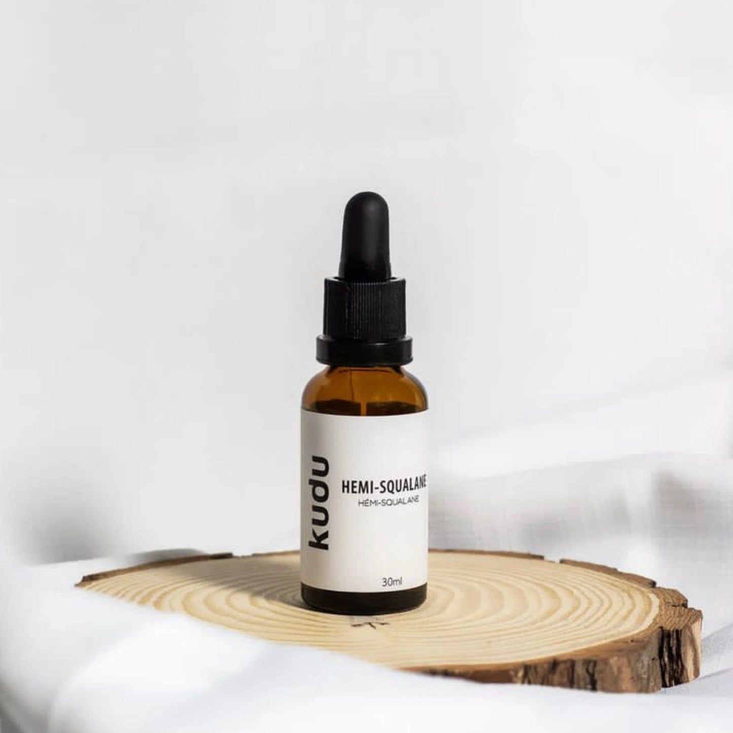 Brown dropper bottle labeled "Kudu Hemi-Squalane" on a wooden slice against a white background. Minimalist and natural skincare theme