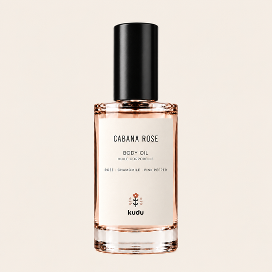 A bottle of Kudu Cabana Rose Body Oil with a black cap and a cream-colored label, set against a solid light beige background.
