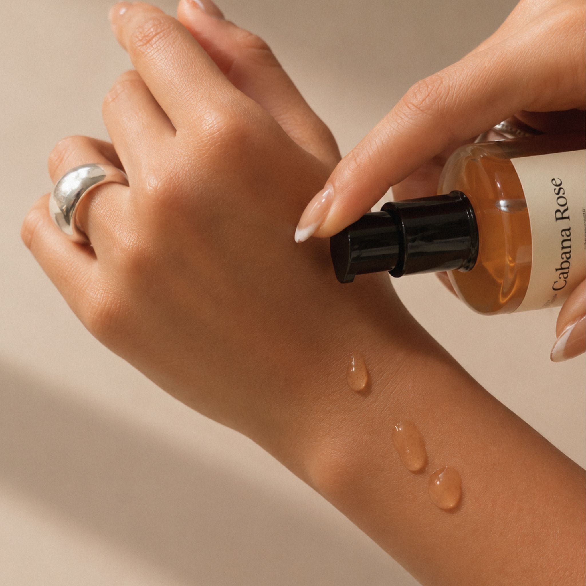 A close-up of a hand receiving a cosmetic product, with another hand holding a bottle labeled 'Cabana Rose' and applying a glossy amber liquid in three drops onto the skin against a warm beige background.