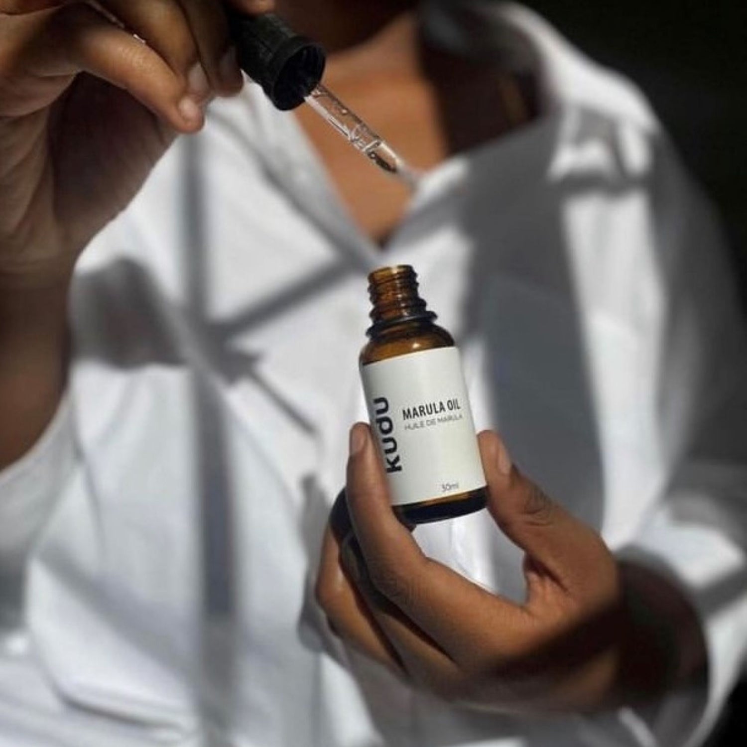 A person in a white shirt holds a dropper above a brown bottle labeled "Kudu Marula Oil" with a white background.