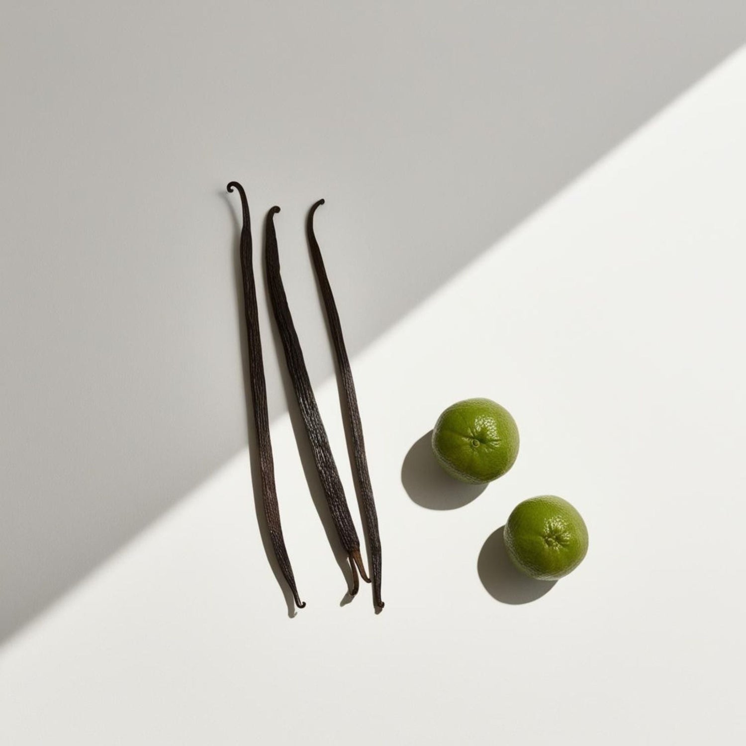 A white surface featuring two green citrus fruits alongside a vanilla pod, evoking a fresh bergamot and vanilla aroma.